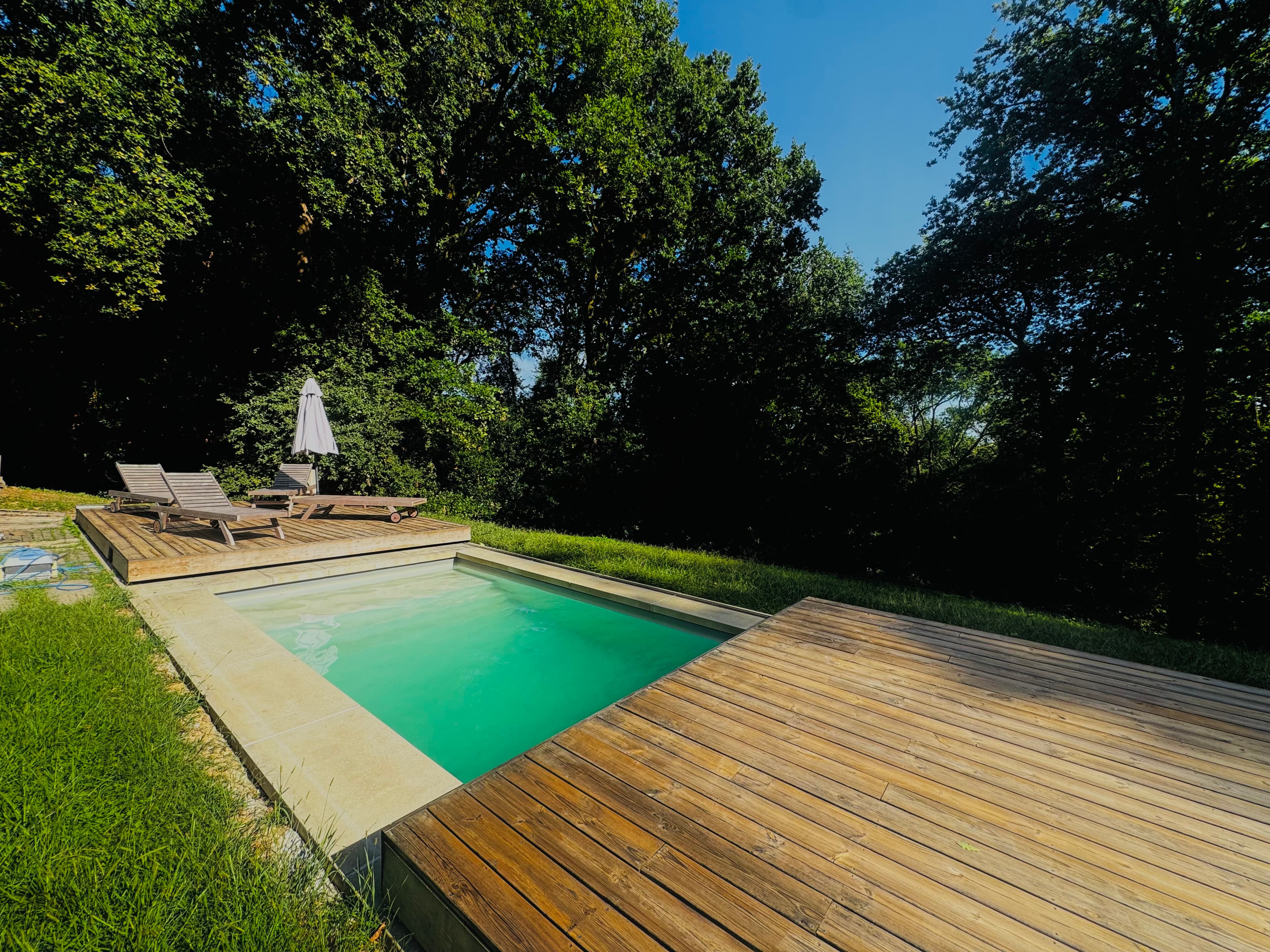Piscine turquoise entourée d'une terrasse en bois et d'arbres verdoyants sous un ciel bleu.