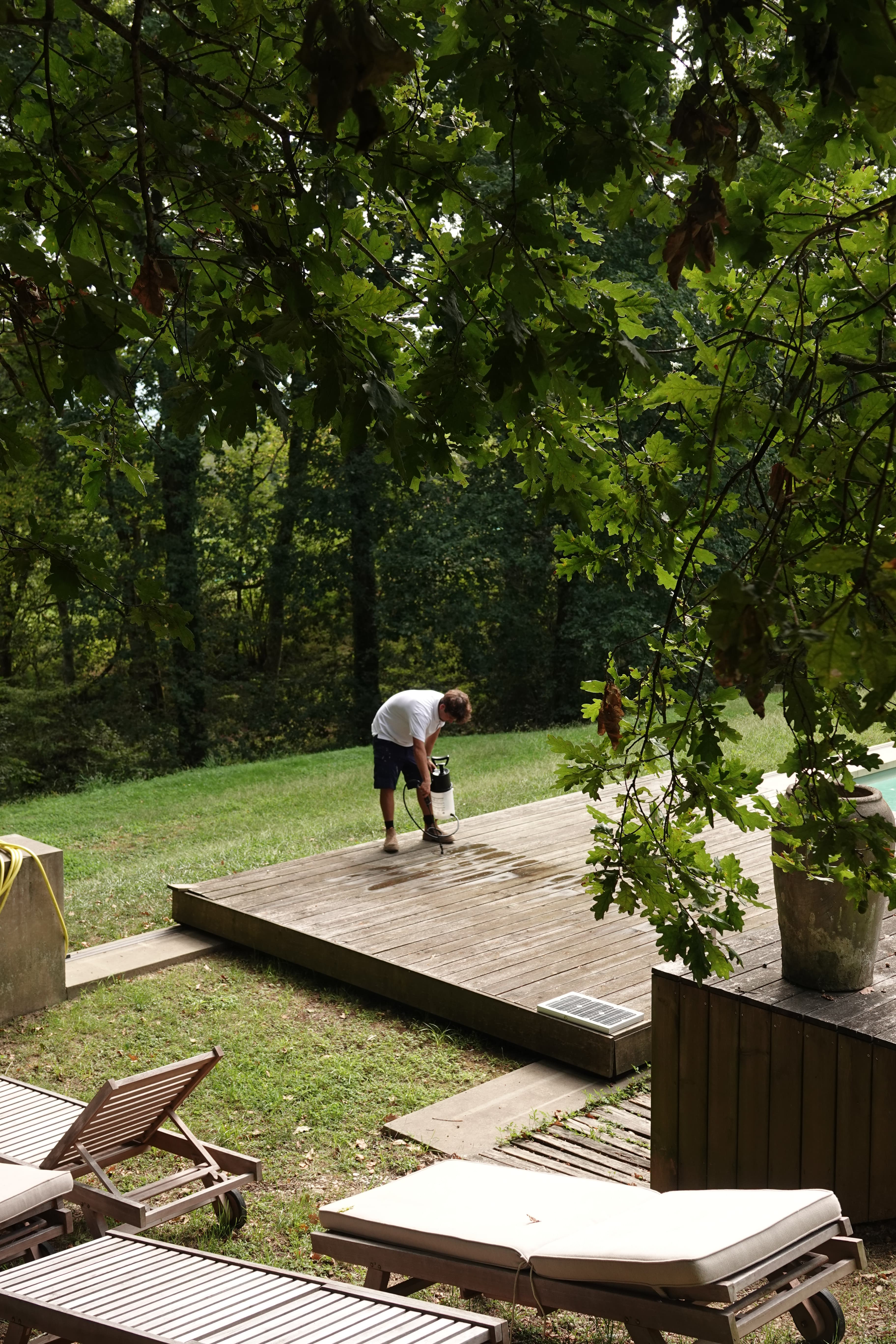 Un homme traite une terrasse en bois avec un pulvérisateur dans un jardin verdoyant.