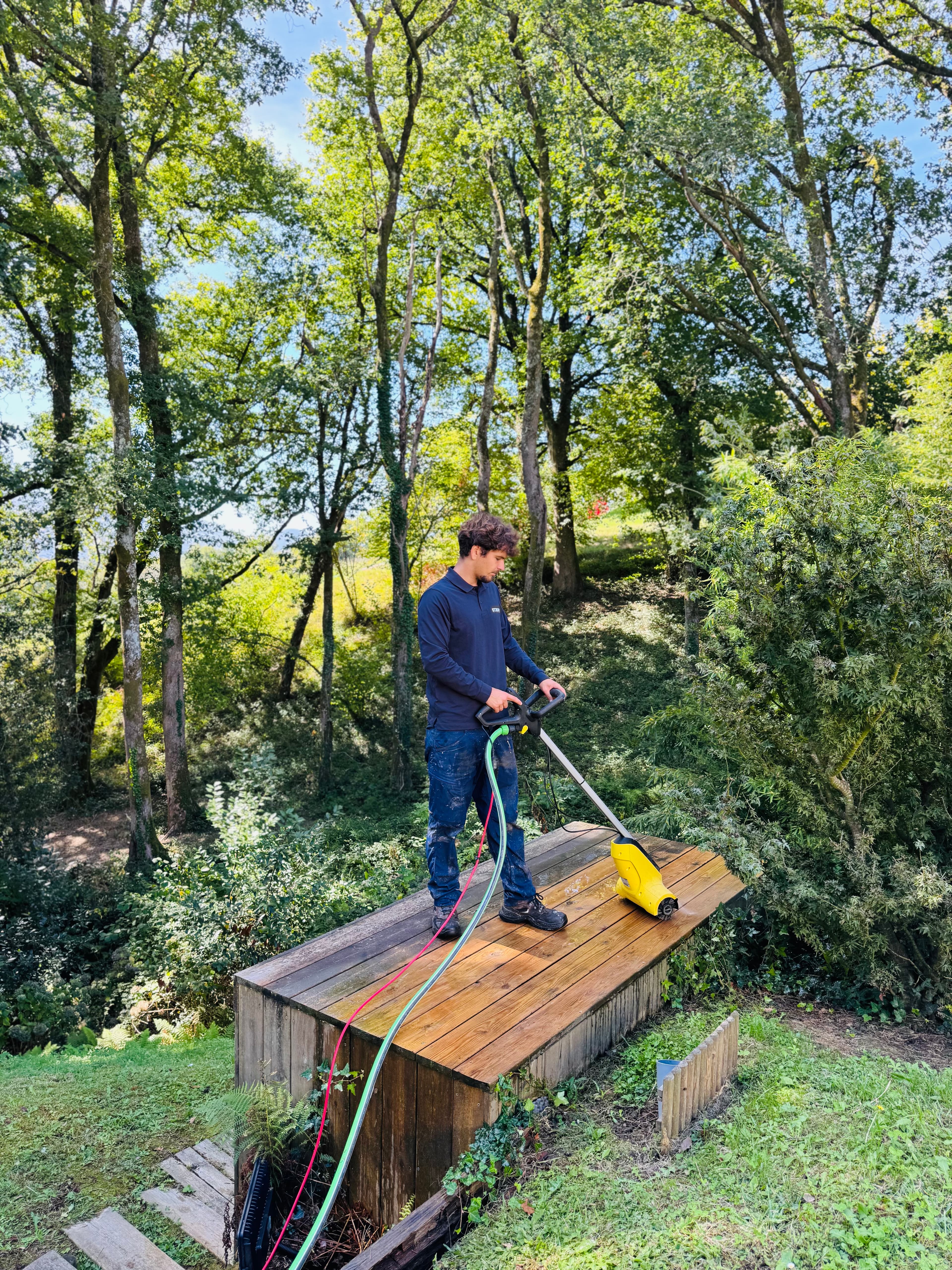 Homme utilisant un nettoyeur de terrasse jaune sur une plateforme en bois entourée d'arbres.